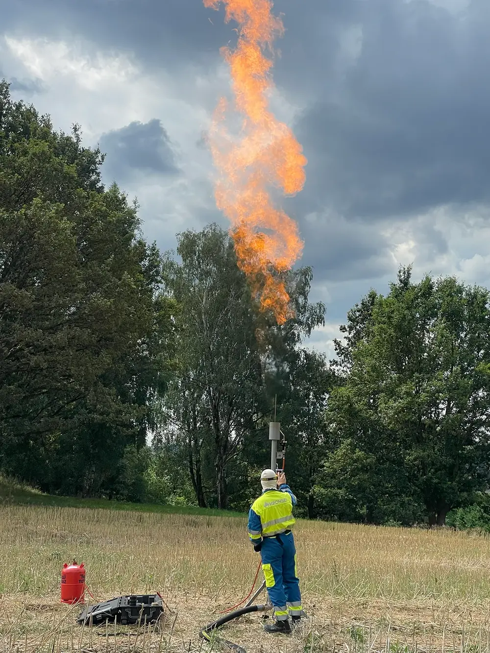 Mitarbeiter mit Helm und Warnkleidung neben Feuerlöscher und Transportkoffer, die Flamme im bewölkten Himmel.