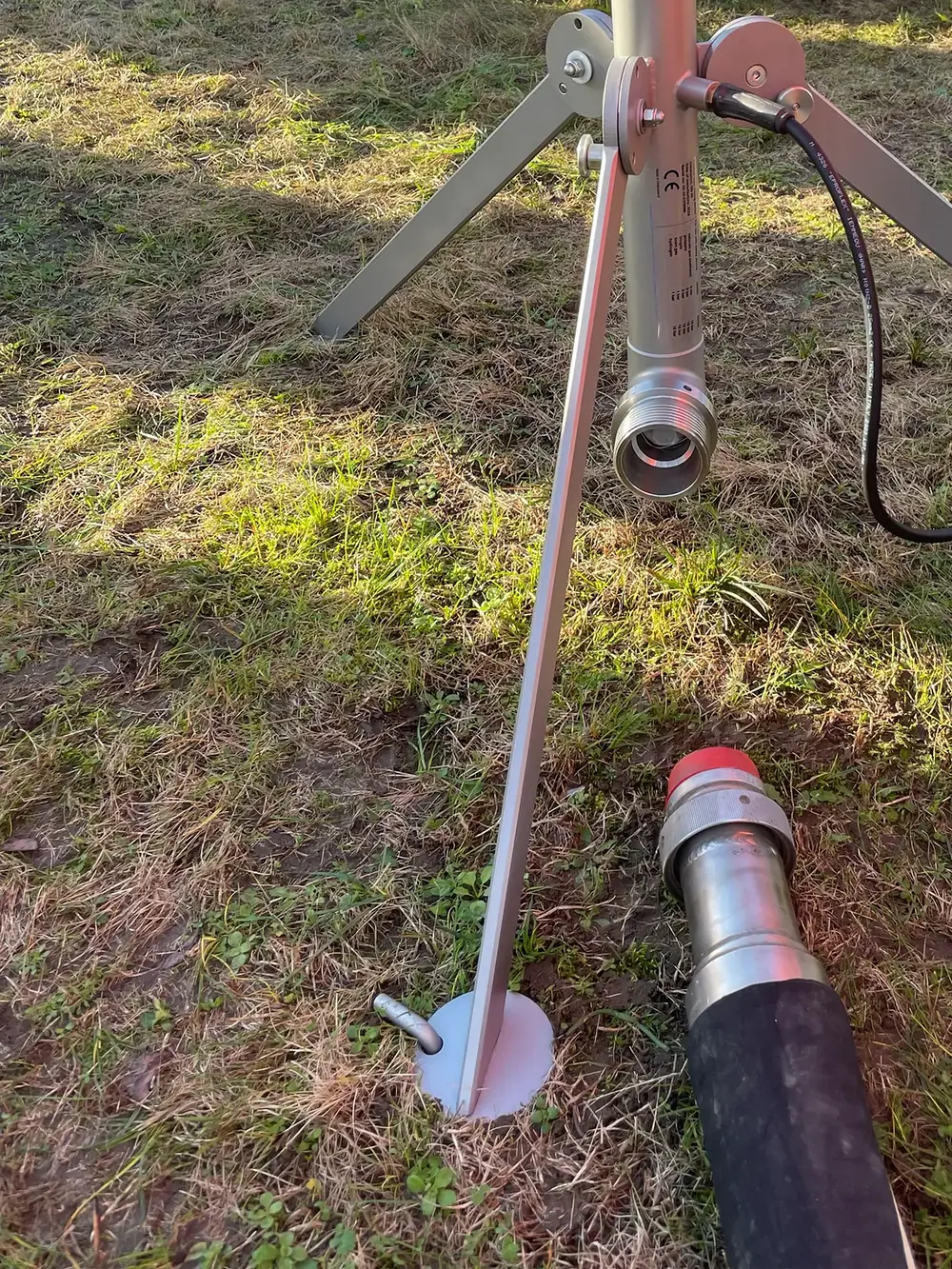 Nahaufnahme des Stativfußes mit Erdspieß im Gras, daneben der angeschlossene Entgasungsschlauch.
