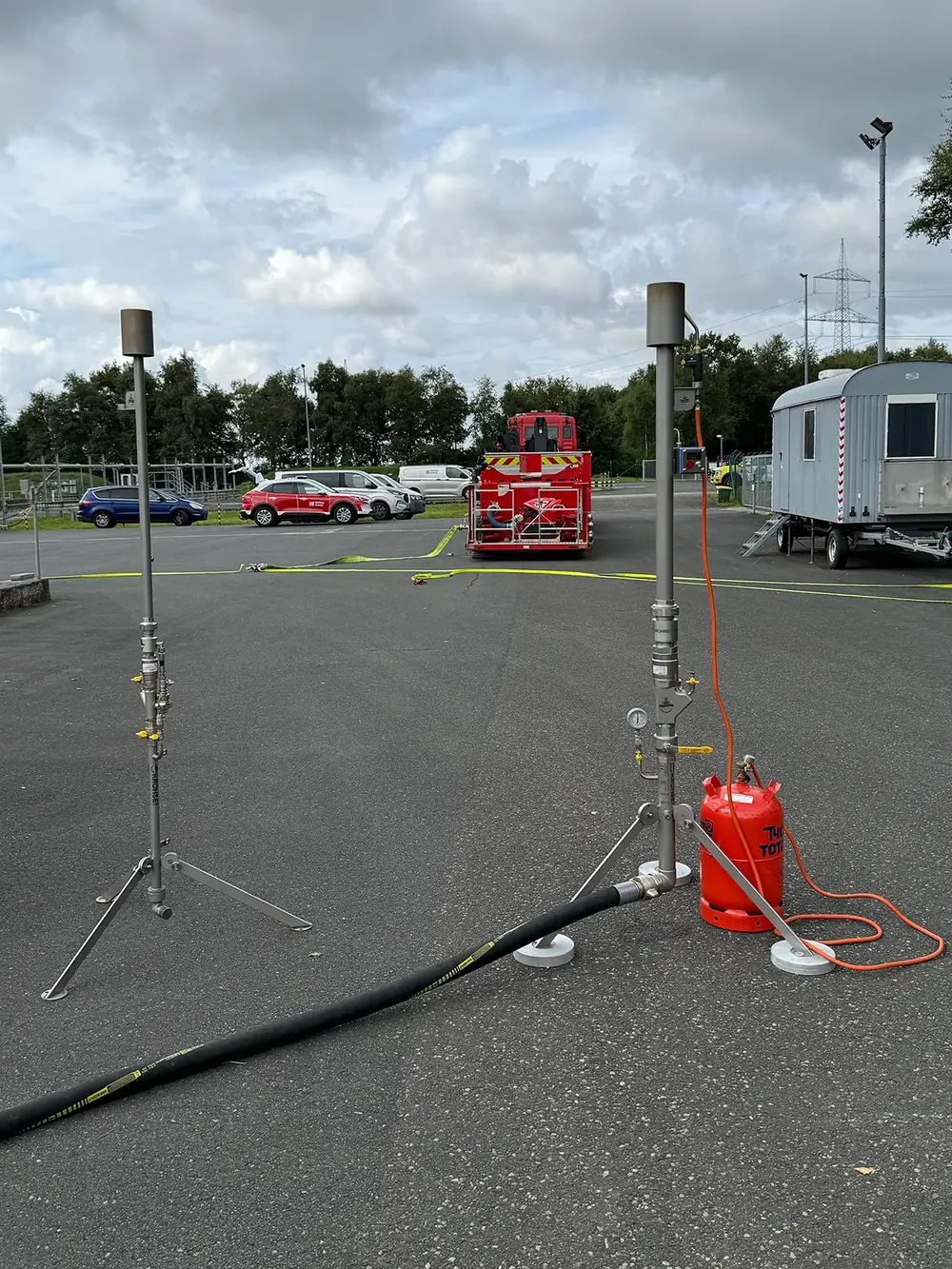 Zwei Gasfackeln nebeneinander auf einer Asphaltfläche, im Hintergrund ein Feuerwehrfahrzeug.