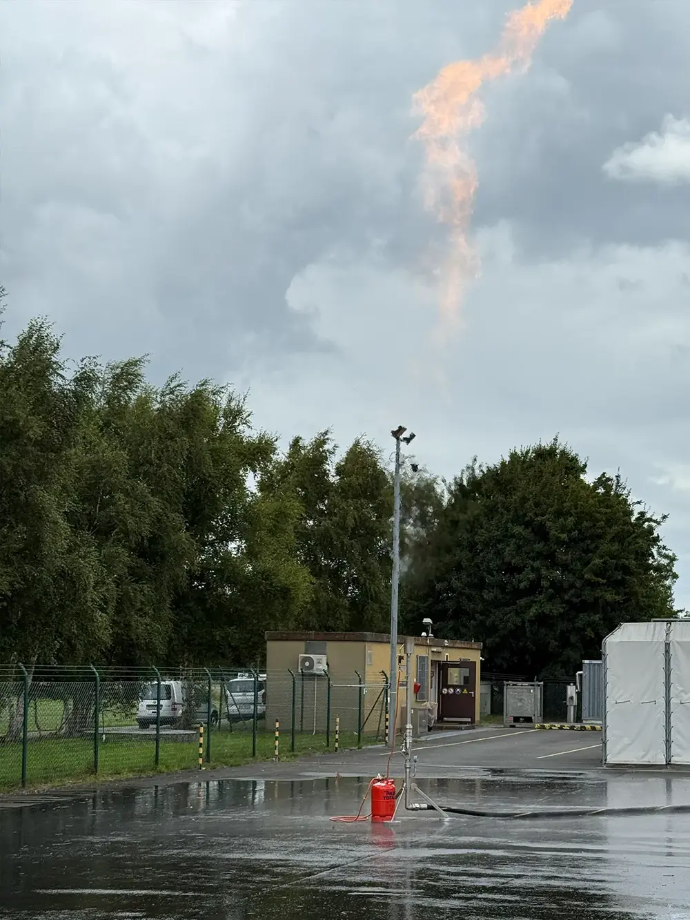 Gasfackel bei Regen auf dem Gelände einer Station, daneben ein Feuerlöscher.