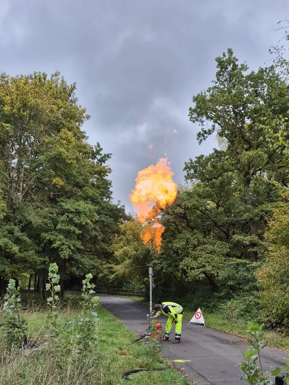 Mitarbeiter arbeitet an der Anschlussstelle am Wegrand, darüber die Flamme über den Baumkronen.